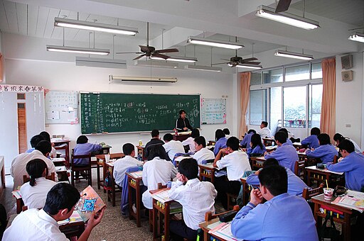 Students in blue or white shirts sitting in rows in classroom facing a green chalkboard
