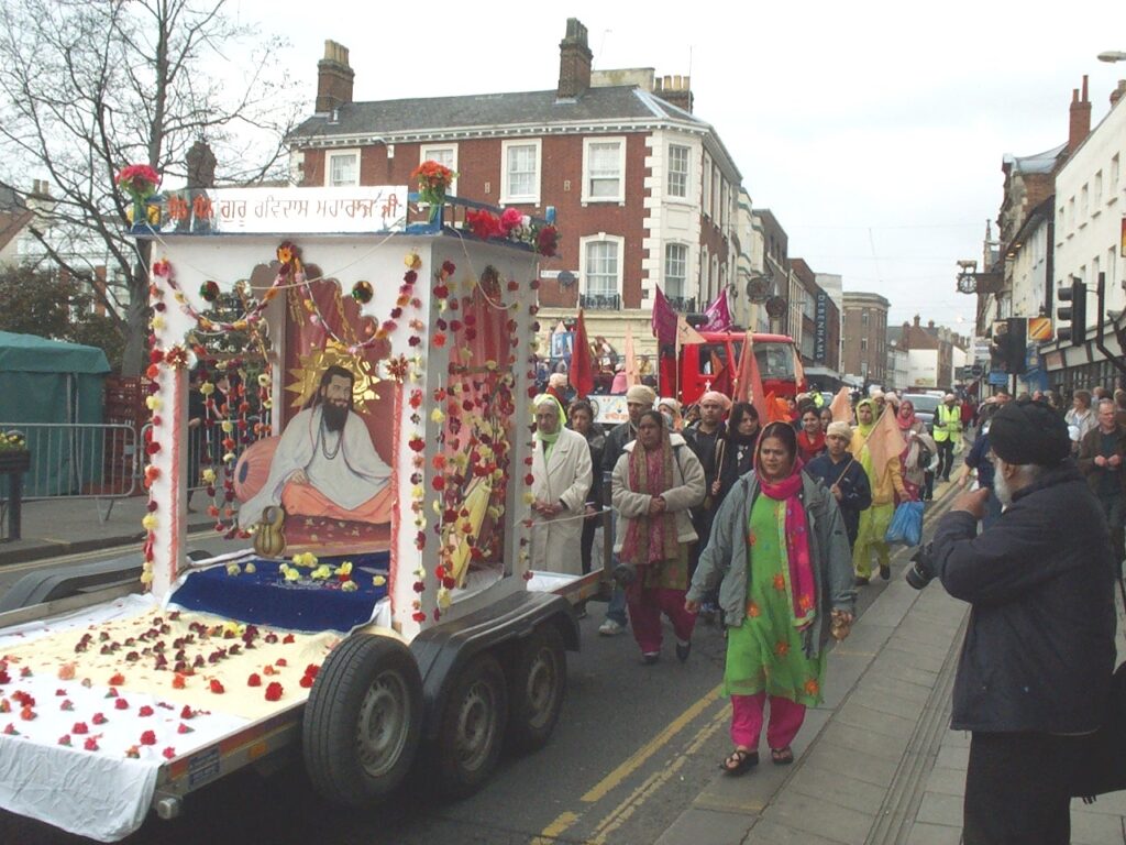 Photo of procession in city street, with people of Indian heritage following a trailer carrying an image of Ravidas in a shrine, surrounded by orange, red, and yellow flowers