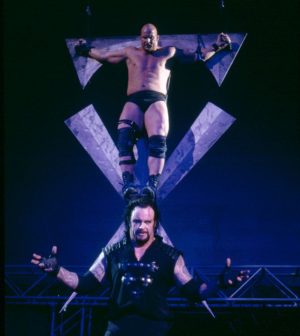 Photograph of Steve Austin hanging from a giant crucifix-like symbol while The Undertaker poses in the foreground.