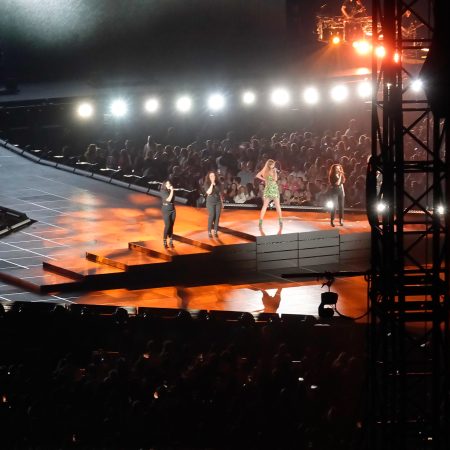 Taylor Swift in green dress on stage with backup singers at Eras concert, fans in background